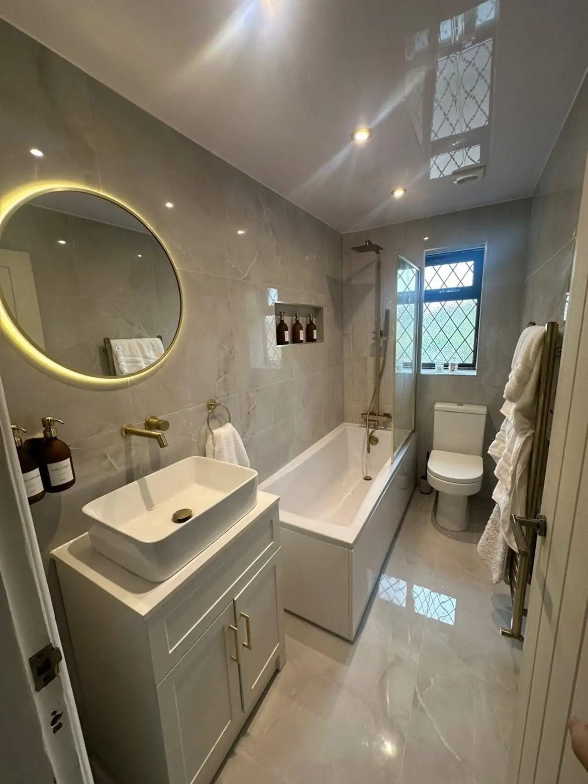 Galley-style bathroom with grey marble-effect tiles, brushed brass fittings, backlit round mirror and bathtub with rainfall shower