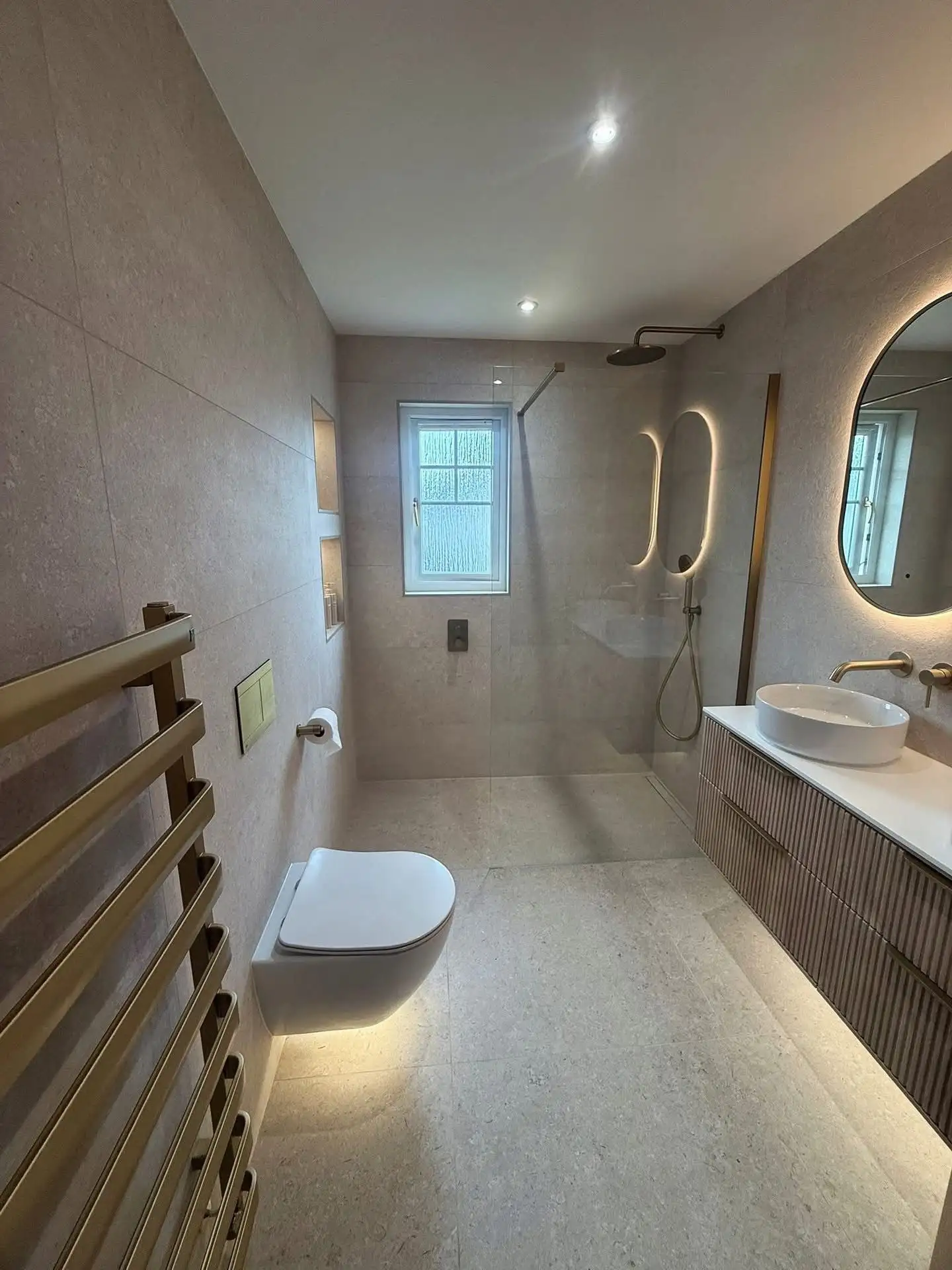Walk-in shower room with beige stone-effect tiles, brushed brass shower fittings, wall-mounted toilet and fluted vanity unit